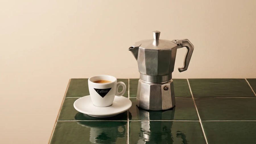 Espressokocher aus Aluminium neben einem Espresso in der Cellini-Tasse auf dunkelgrünen Fliesen