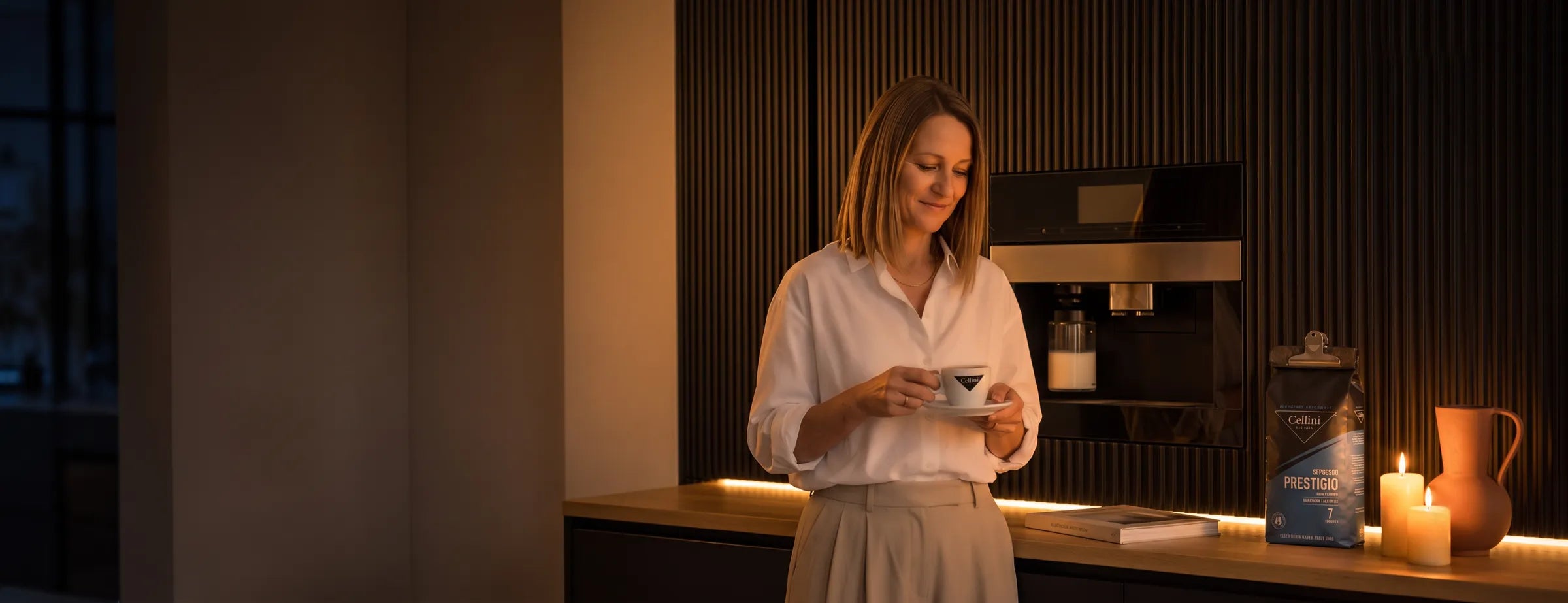 Frau steht am Morgen vor dem Einbau-Vollautomaten in der Küche und schaut auf ihren Milchkaffee in der Cellini-Tasse, daneben der mit der Cellini-Klammer verschlossene Beutel Espresso Prestigio 100 % Arabica Bohnen 1 kg bei Kerzenlicht