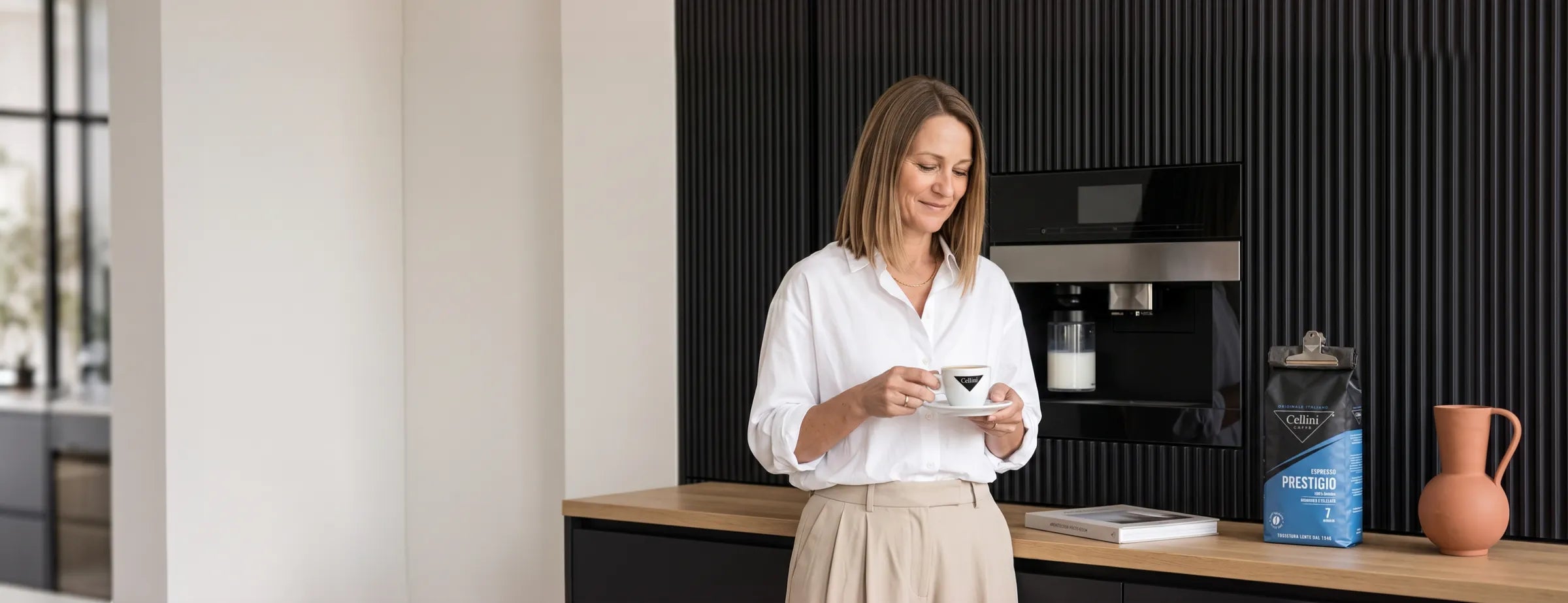Frau steht am Morgen vor dem Einbau-Vollautomaten in der Küche und schaut auf ihren Milchkaffee in der Cellini-Tasse, daneben der mit der Cellini-Klammer verschlossene Beutel Espresso Prestigio 100 % Arabica Bohnen 1 kg