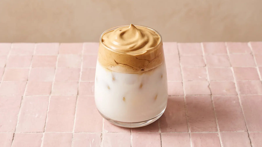 Dalgona Coffee: luftiger Kaffeeschaum auf kalter Milch mit Eis in einem mundgeblasenen Glas auf rosé Zellige-Fliesen