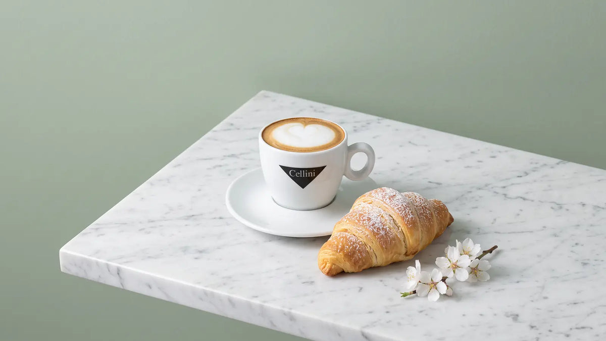 Muttertags-Frühstück: Cappuccino in der Cellini-Tasse, ein Cornetto mit Puderzucker und ein Mandelblütenzweig auf weißem Marmor