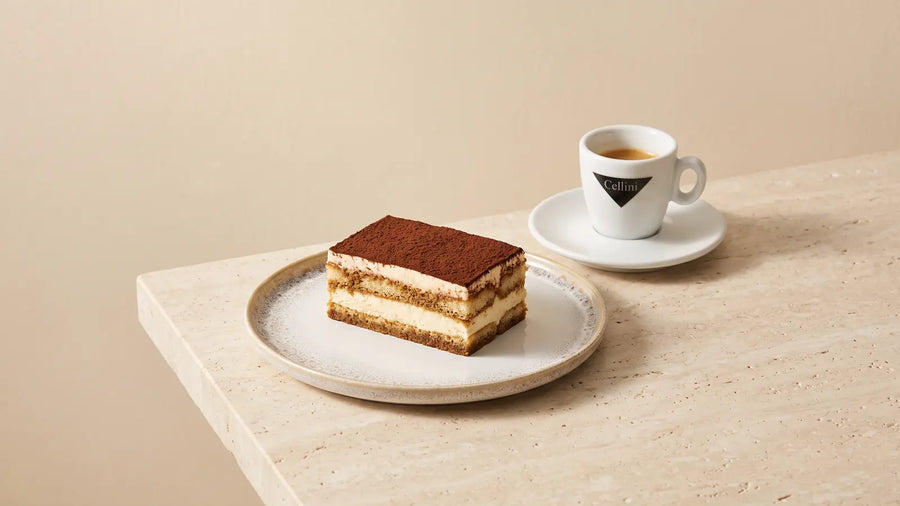 Ein Stück Tiramisu mit Schichten aus Löffelbiskuits, Mascarponecreme und Kakao auf einem Keramikteller, daneben ein Espresso in der Cellini-Tasse