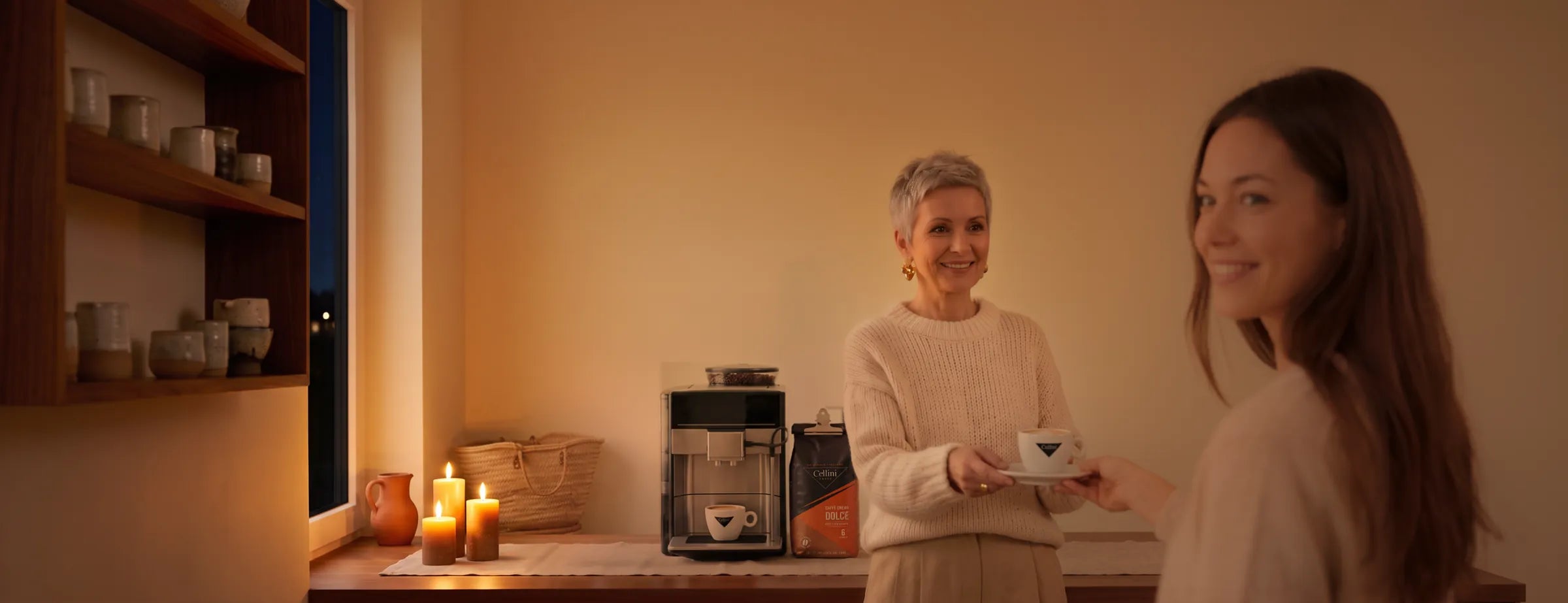 Frau reicht am Sonntagnachmittag einer Freundin einen Caffè Crema in der Cellini-Cappuccinotasse, daneben Kaffeevollautomat mit zweiter Tasse unter dem Auslauf und der mit der Klammer verschlossene Beutel Cellini Caffè Crema Dolce Bohnen 1 kg bei Kerzenlicht