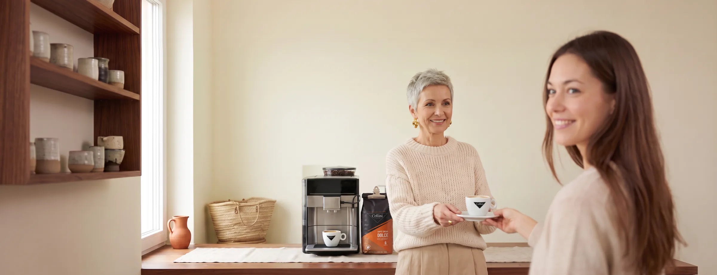 Frau reicht am Sonntagnachmittag einer Freundin einen Caffè Crema in der Cellini-Cappuccinotasse, daneben Kaffeevollautomat mit zweiter Tasse unter dem Auslauf und der mit der Klammer verschlossene Beutel Cellini Caffè Crema Dolce Bohnen 1 kg